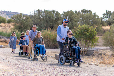 A group of people on the beach. Three are standing, one has a prosthetic leg and the other two are on beach wheelchairs. 
