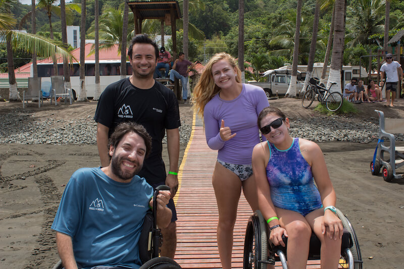 A group of people on the beach. Three are standing, one has a prosthetic leg and the other two are on beach wheelchairs. 