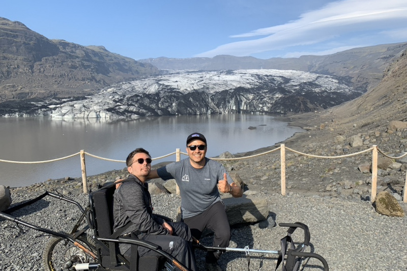 Two people, including a person in a wheelchair, pose for a photo in front of the geyser.