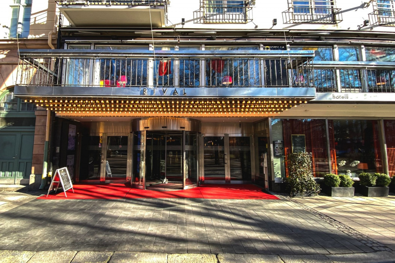 The outside view of Hotel Rival shows carpet flooring, a revolving door, and two french glass doors on each side