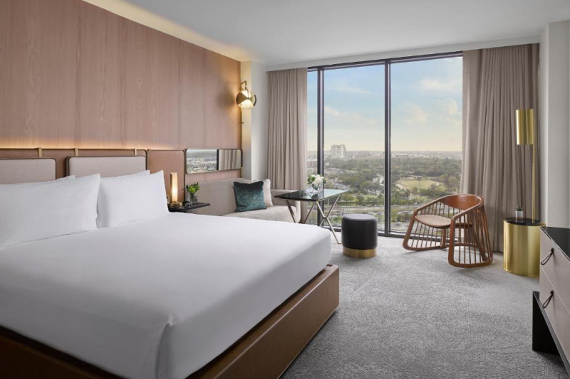 This room has a king-sized bed with a floor-to-ceiling window at one end. There is a dresser opposite the bed as well as a sofa, a chair, a small table, and a footstool.