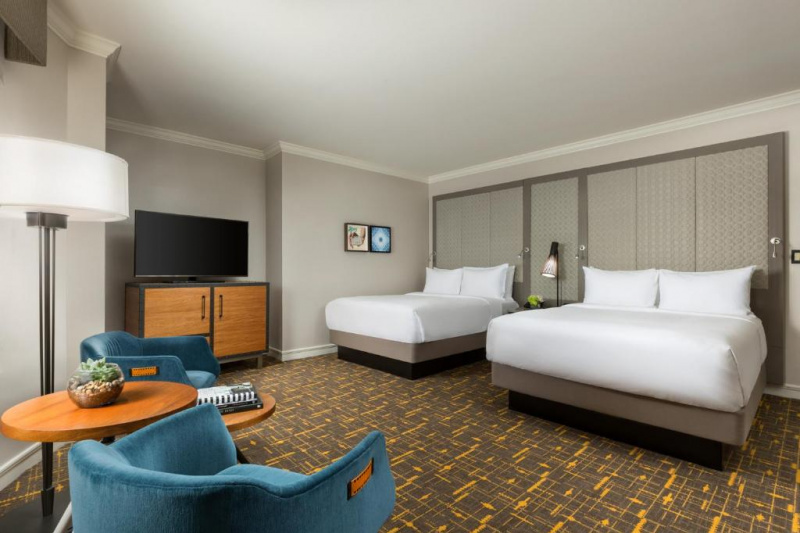 This accessible junior suite two queen has two queen-sized beds each with a padded headboard. There is a nightstand between them, with a reading lamp on it. To one side is a sideboard with a TV on top. Opposite the beds is a seating area with two armchairs, a low table and a floorstanding lamp.