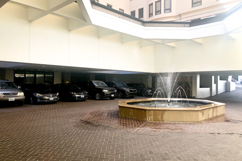 There are 6 cars parked in the hotel's semi underground parking garage. The large brick-paved area has a fountain in the middle.