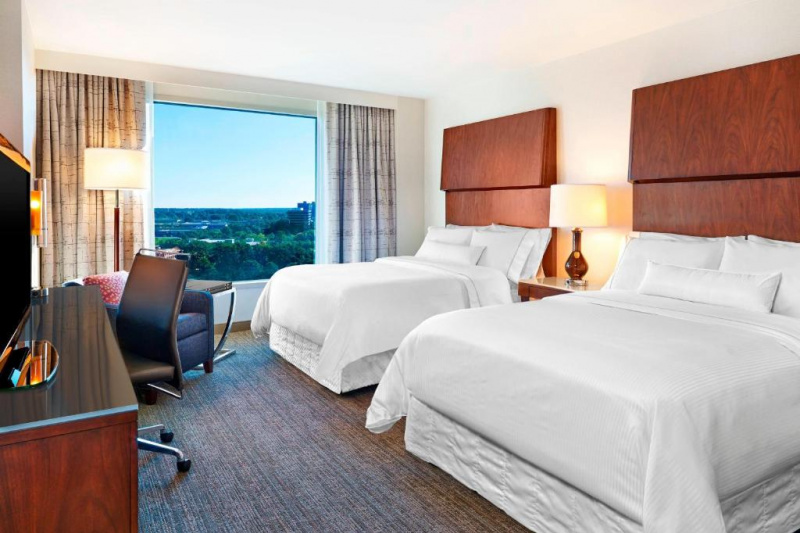 This accessible 2 queen guest room has 2 queen-sized beds, a nightstand, and a lamp. Opposite the bed, there is a flat-screen TV, an armchair and a table and a chair with wheels.