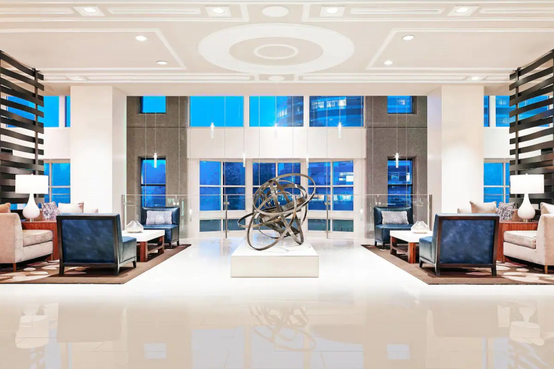The accessible well-lit hotel lobby has a spacious seating area with assorted tables and chairs. In the center of the lobby there is metal sculpture.