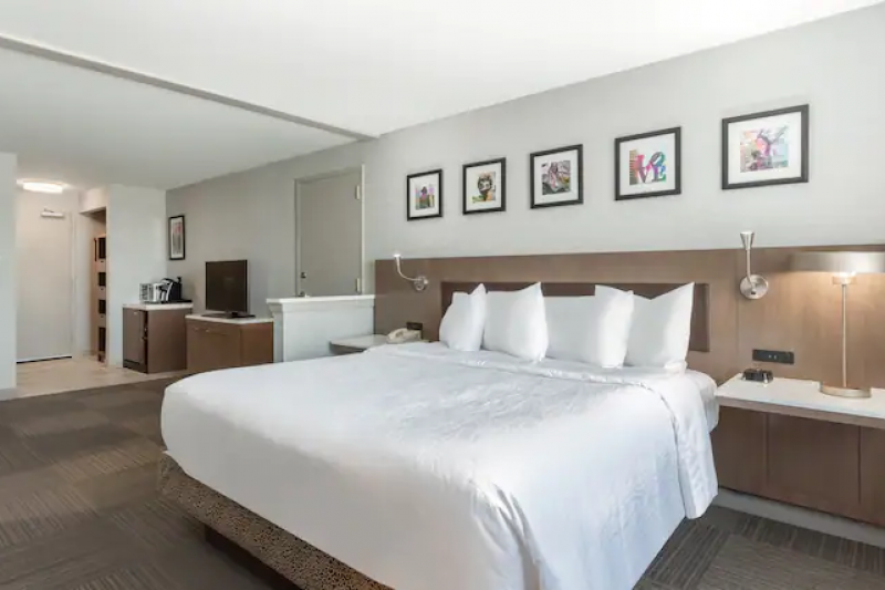 This guest room has a king-sized bed. The wooden headboard has a nightstand built-in on either side of the bed. There are wall-mounted lamps above the nightstands. Further into the room is a chest of drawers with a TV on top and another chest of drawers with a coffee maker.