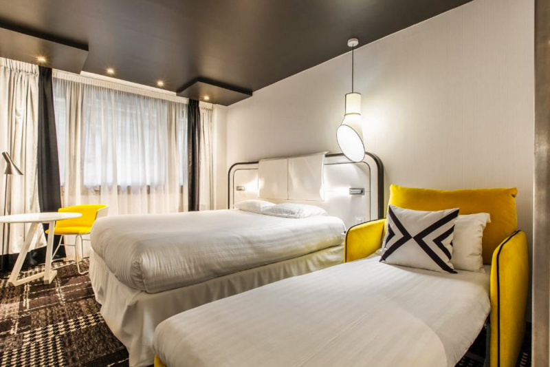 This deluxe triple room has a double bed and an additional single bed. There is a large window to one side and a small coffee table with two chairs. There is a floor standing lamp next to the table.