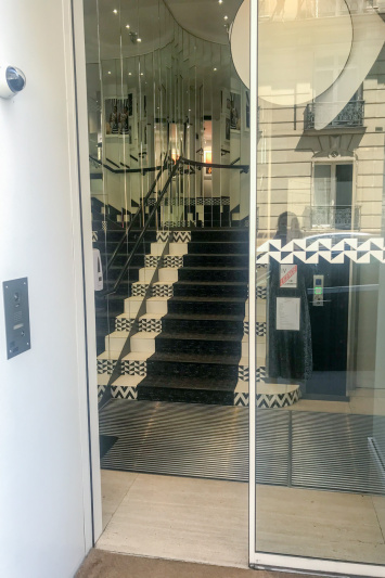 The entrance to the hotel is through a glass door. There is an intercom to one side. Inside, there is a black and white patterned staircase with mirrored walls.