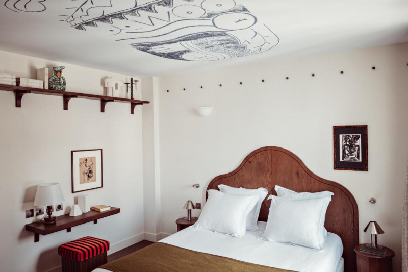 The executive room has a double bed with a wooden headboard. There are nightstands with reading lamps on either side of the bed. There is an additional shelf with a stool and a high shelf with ornaments.