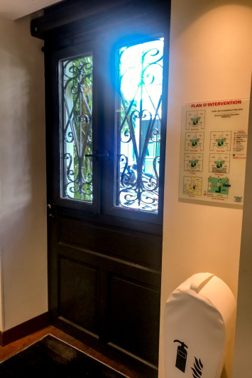 The entrance door is shown from inside the lobby. It has wrought iron work on the two top glass panels. There is a fire extinguisher and a plan of the hotel to the side of it.
