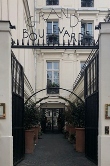 The entrance path to the hotel is from the sidewalk. There is a set of tall, metal gates that open onto a step-free path. On either side of the path are large pot plants.