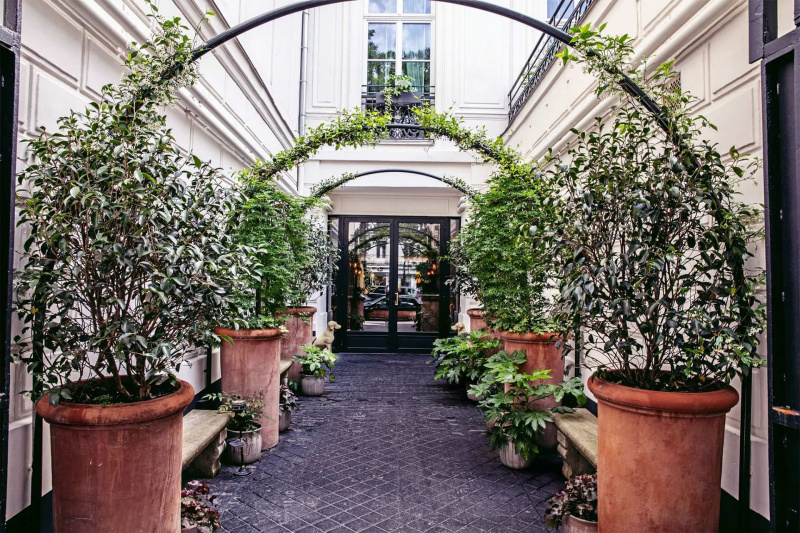 The entrance path to the hotel is lined with large pot plants. Between the pots are stone benches. There is a curved plant canopy above.