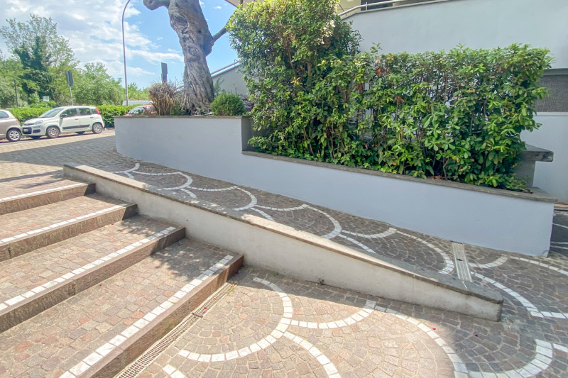 This outdoor cobble stoned area has steps with a ramp at the side. They lead to the road. There is a planter with large plants to the side of the ramp.