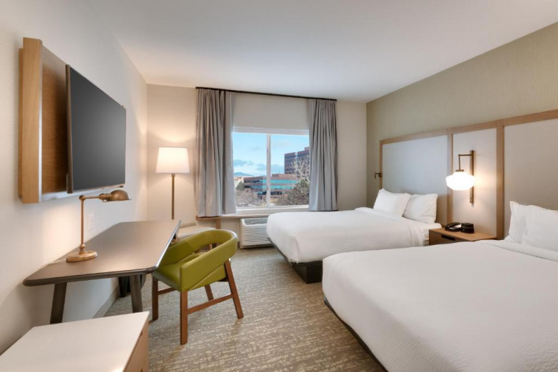 Suite showing 2 queen-sized beds with white linen, wall-mounted TV, desk and chair, under window air-conditioner, lamps and city view.