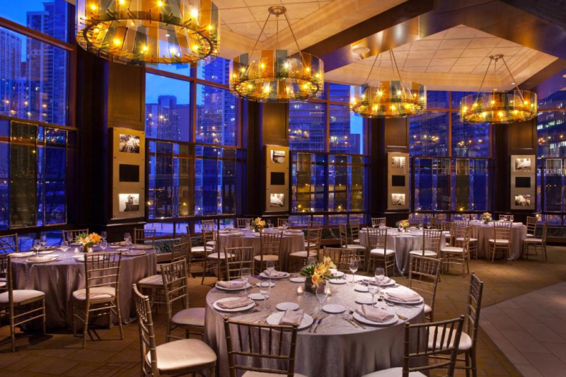 Spacious event venue with circular tables for 8 with moveable chairs and chandeliers overhead. Double height windows provide a cityscape view..