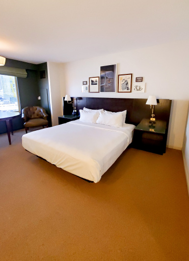 The accessible king has a king-sized bed with white linen, nightstands, wall-mounted reading lamps and artwork, with a wooden table and comfortable armchair by the window.
