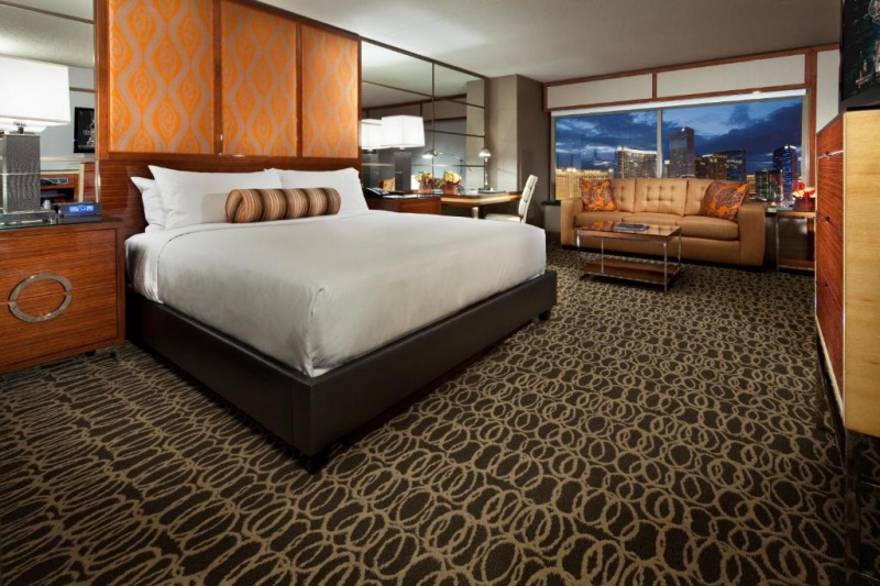 The Grand king room has a king-sized bed with a tall brocade headboard and a mirrored wall. There are consoles with lamps, a radio and a telephone, a leather sofa, a coffee table, and a desk and chair.