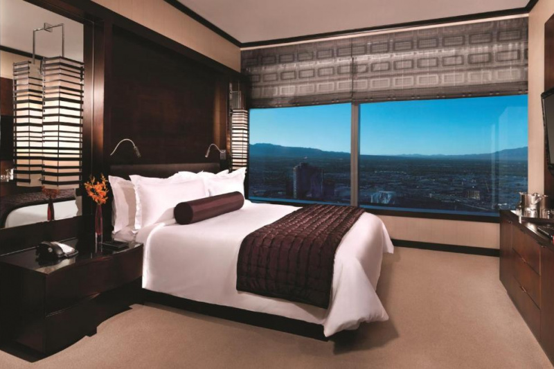 The basic suite has a king-sized bed with white linen and large pillows. There are nightstands on either side of the bed, with hanging lamps above them. On one nightstand is a push-button phone. Opposite the bed is a cabinet with a wall mounted TV above. There is a huge window overlooking the city.