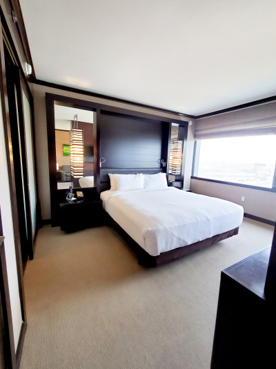 The basic suite has a king-sized bed with a nightstand on each side. On one nightstand there is a telephone. Behind each nightstand there is a mirror with a hanging wall lamp. The bedroom has a large window and low wall sockets.