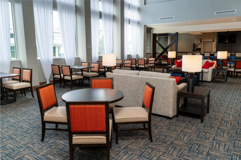 The spacious carpeted seating area has assorted tables, chairs, lamps, and cushions. There are big windows with long with curtains.