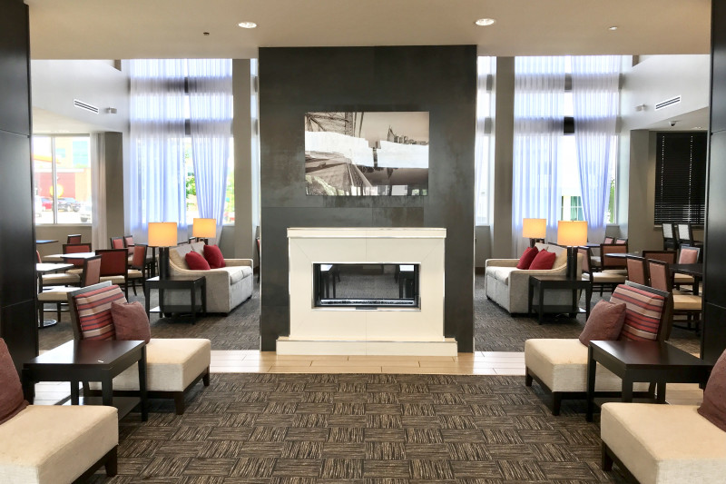 The spacious hotel lobby has assorted tables and chairs, lamps, cushions, an electric fireplace and artwork on the wall.