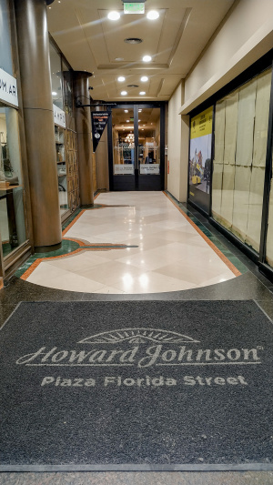 The well-lit hotel entrance has two glass doors. The entrance is lined with shops.