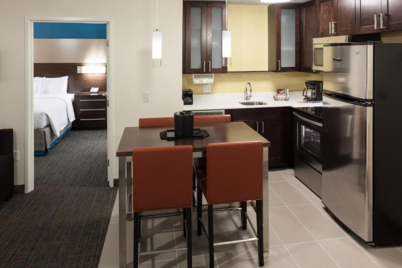 The 1 bedroom suite has a kitchen with oven, microwave, refrigerator, and a dining table and 4 chairs. There is a king-sized bed with a nightstand and a mounted light in the bedroom.
