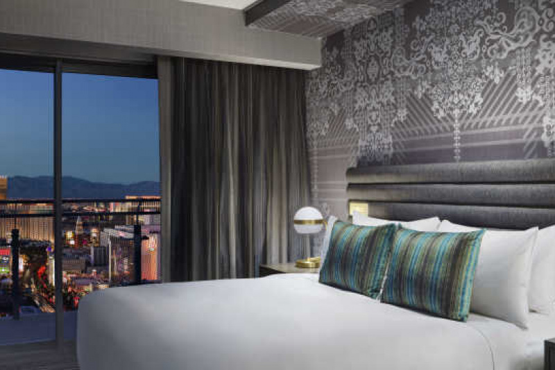 The terrace one bedroom suite fountain view has a king-size bed with nightstands on either side. There are lamps on the nightstands. At the far end of the room is a large floor to ceiling window.