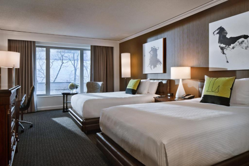 The guest room has a king-sized bed with nightstands o either side of it. There is a window at one side of the room with a door leading to a balcony. Opposite the bed is a chest of drawers with a TV on top.