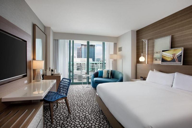 The room has a king-sized bed with nightstands. There is a wall-mounted TV, a workstation with movable chair, and a small sofa with cushions.
