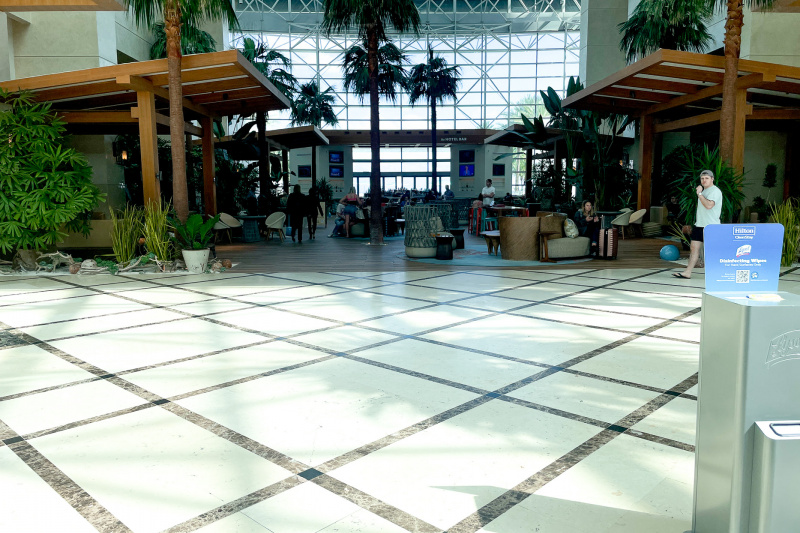 The spacious hotel lobby has a central area filled with palm trees and numerous seating areas with assorted tables and chairs. The hotel bar is at the far end.