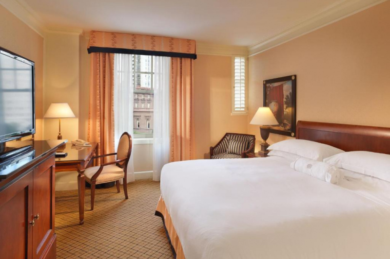 This Standard guest room has 1 king-sized bed. There is an armchair in the corner. Opposite the bed is a desk, chair, and a tall chest of drawers with a TV on top. The room is carpeted and spacious.