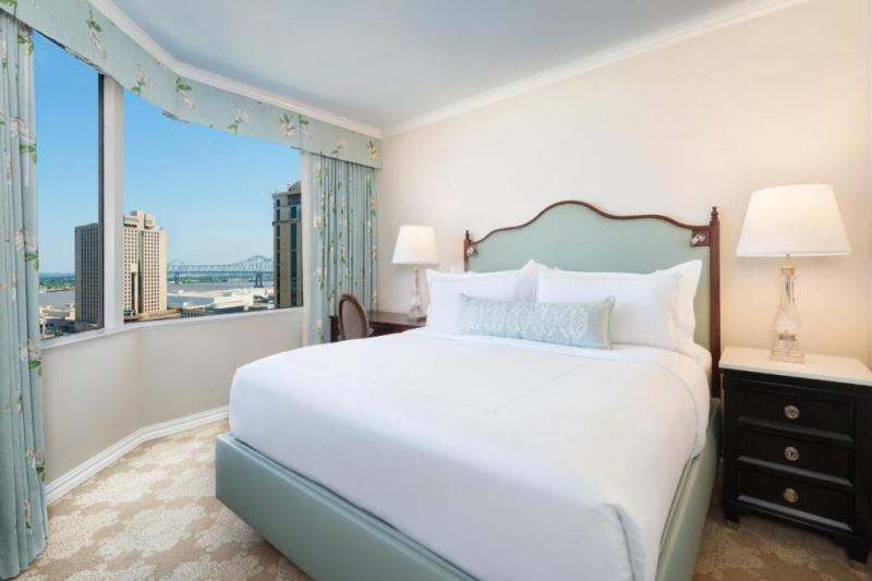 The hotel guest room has a king-sized bed with white linens. On one side of the bed, there is a nightstand with a lamp, on the other, there is a small table with a lamp and chair.
