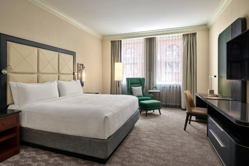 This guest room has 1 king-sized bed, nightstands, a high-back armchair, a footstool, and a wall-mounted TV. There is also a desk with a small chair and a lamp.