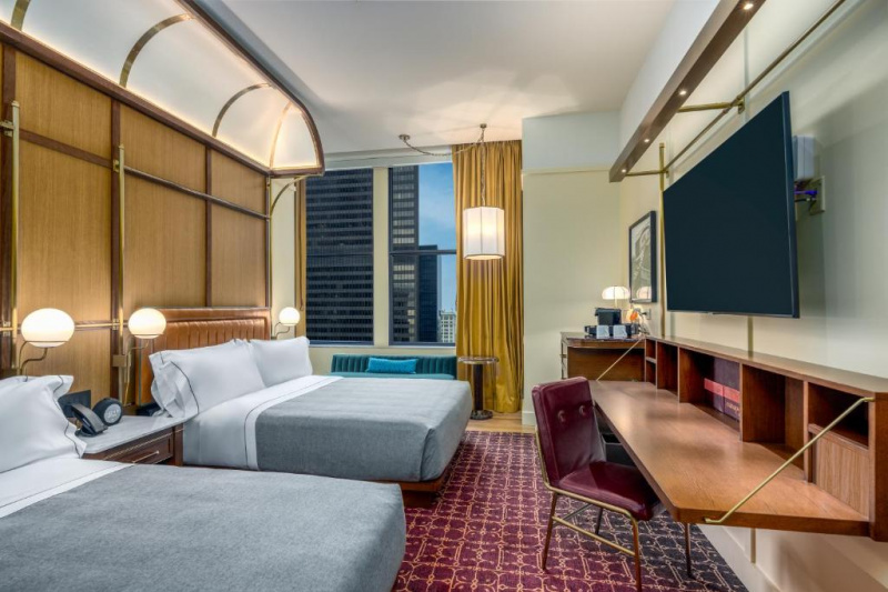 The spacious carpeted room has 2 queen-sized beds, a central nightstand with wall mounted reading lamps. There is a large wall-mounted TV, a foldaway workstation with a movable chair, a chest of drawers, and a low-backed sofa under the window.