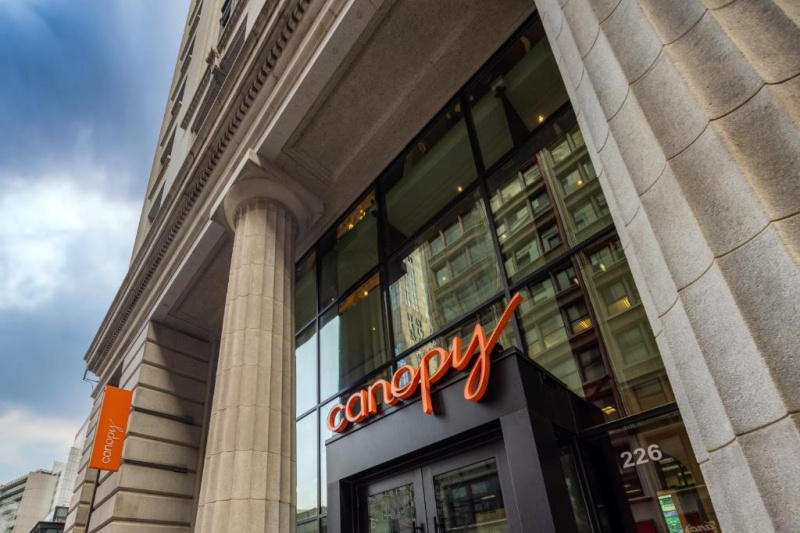 The hotel has a modern entrance with double glass doors under a tall older-style stone column portico. The hotel's name is written immediately above the modern entrance.