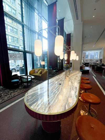 The spacious hotel lounge has a staffed serving counter. There are assorted low tables, sofas, movable chairs and stools. In the center of the room there is an oval counter with bar stools.