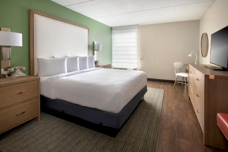 The room has a king-sized bed, 2 nightstands with a telephone and wall-mounted lamps. There is a chest of drawers, a flat screen TV, a workstation and a movable chair.