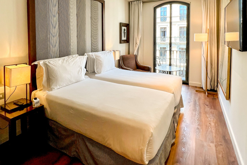 The room has 2 twin beds pushed together, and nightstands with lamps on either side. There is a wall-mounted TV, a small armchair with a round table and a standing lamp.