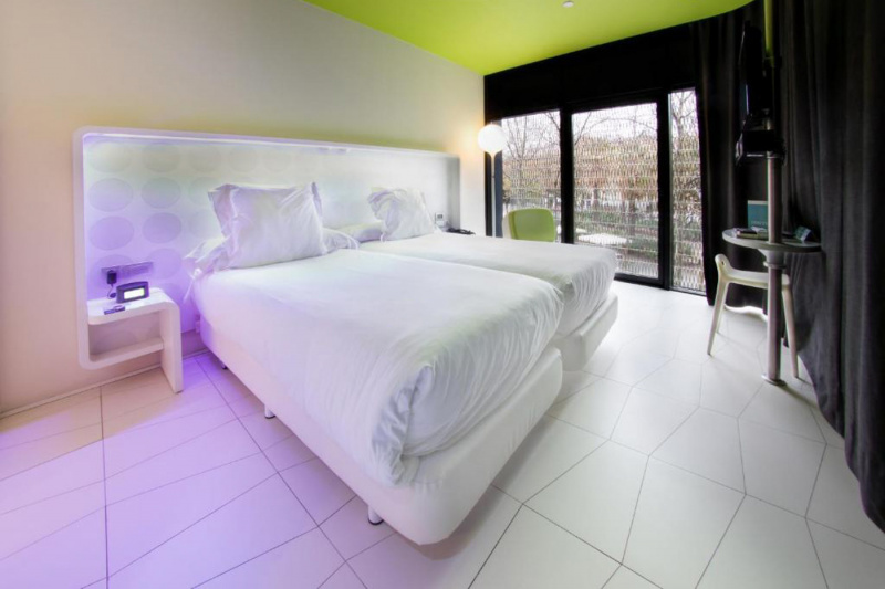 The room has 2 twin beds with white linen pushed together, nightstands with plug sockets, a chair, a small workstation and a stool.