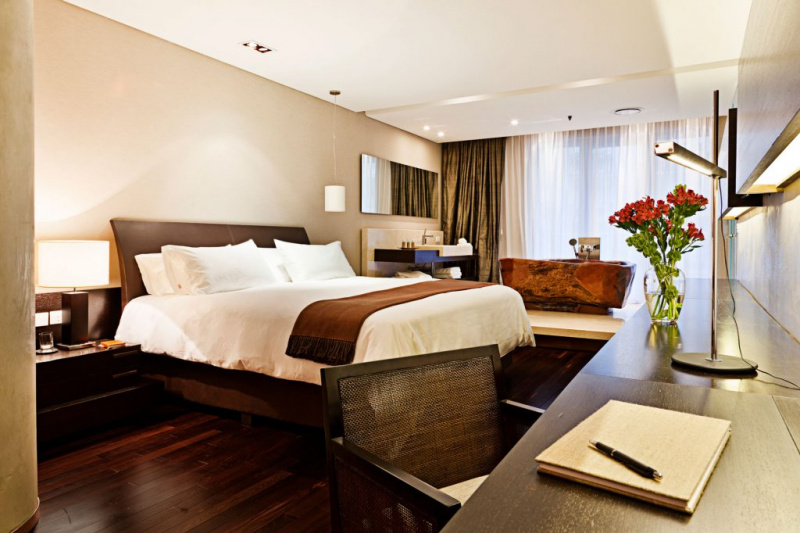 The room has a king-sized bed, with a nightstand and a lamp. There is a desk and chair opposite the bed. The ensuite bathroom has a step leading to a free-standing wooden bathtub and sink unit with shelves