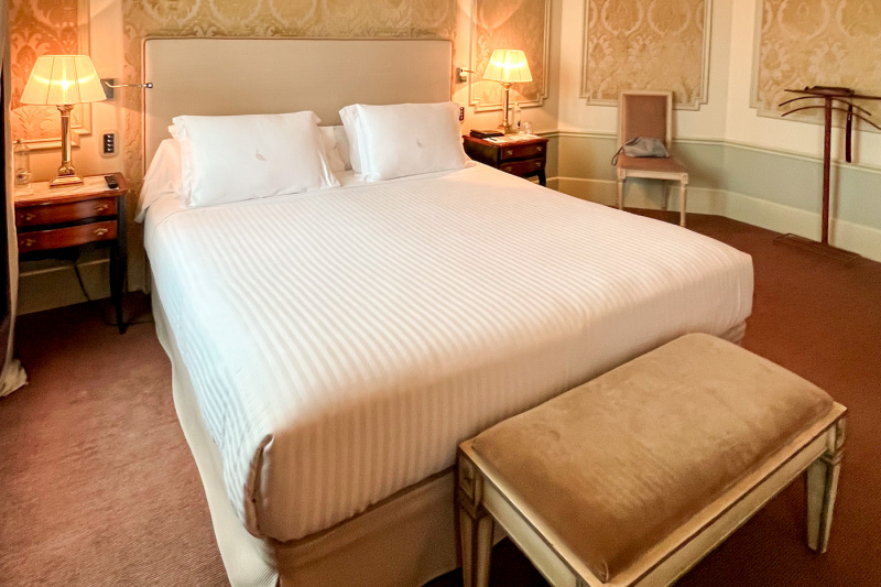 The spacious room has a king-sized bed with a plush bed bench at the end. There a nightstands on either side of the bed, with lamps on them.