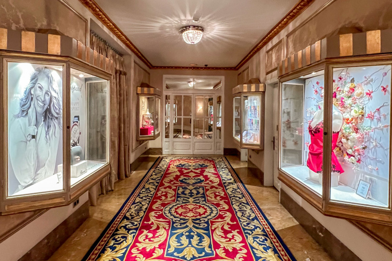 The area has an ornate rug on the corridor floor, leading to some double doors. The corridor is lined with small glass cabinets displaying perfume and lingerie.