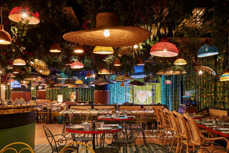 The colorful dining area has wicker lights hanging from the ceiling. There are a number of movable tables and chairs in the room with a food counter to one side.