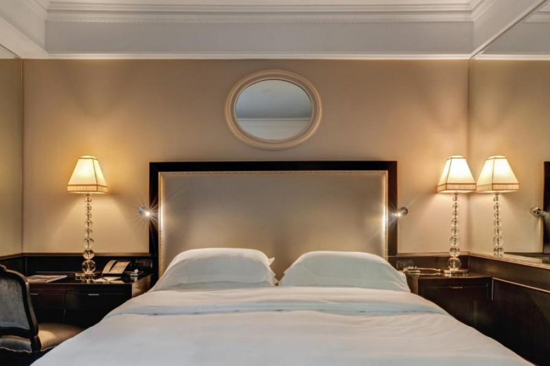 The room has a king-sized bed with a nightstand on one side. There is a desk and chair on the other side of the bed. There are table lamps on the nightstand and the desk.