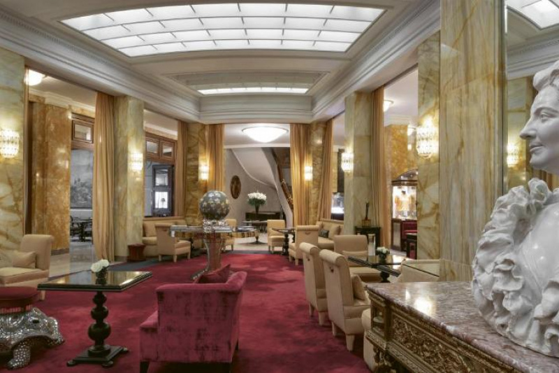 The long, ornate, seating area is carpeted. There are heavy, movable chairs and tables in the room, with space between the seating areas.