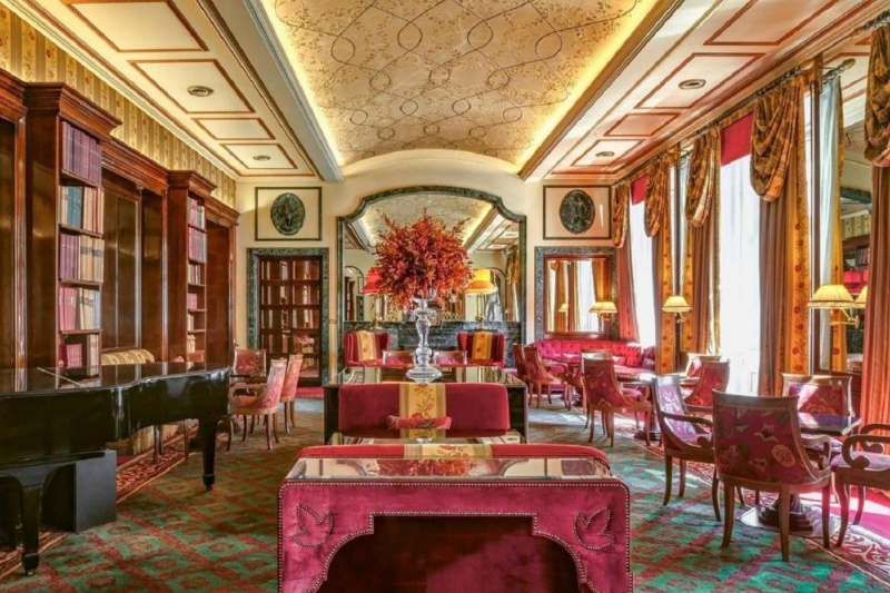 The ornate piano room has sets of heavy, movable chairs and tables. There is space between the sets of tables and chairs.