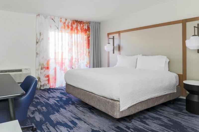 This spacious room has a king-sized bed with nightstands on either side. Above the nightstands there are wall-mounted hanging lamps. There is also a workstation and a movable tub chair.