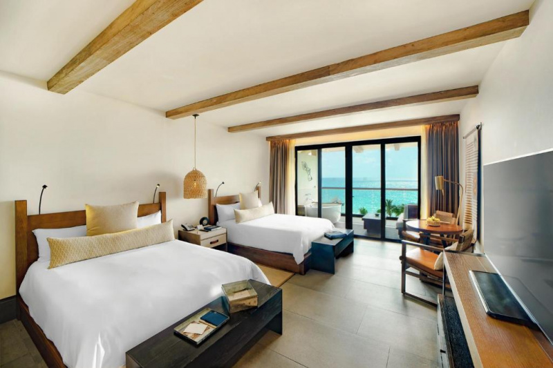 The spacious room has 2 queen-sized beds with a central nightstand. There is a table with 2 chairs, a TV, and wooden benches at the foot of each bed. Sliding doors lead to a balcony.