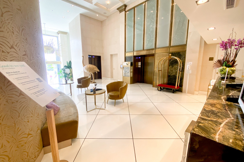 View of the spacious, step-free reception area from the check-in desk. There are 2 small tables and movable chairs. A gold ostrich statue stands next to an elevator. There is a second elevator on another wall next to a luggage trolley.